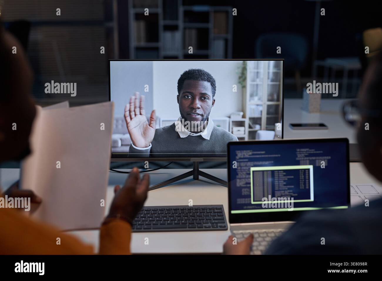 Black male and mixed-race female programmers sitting at table having video call with Black male colleague discussing computer program working late ... Stock Photo