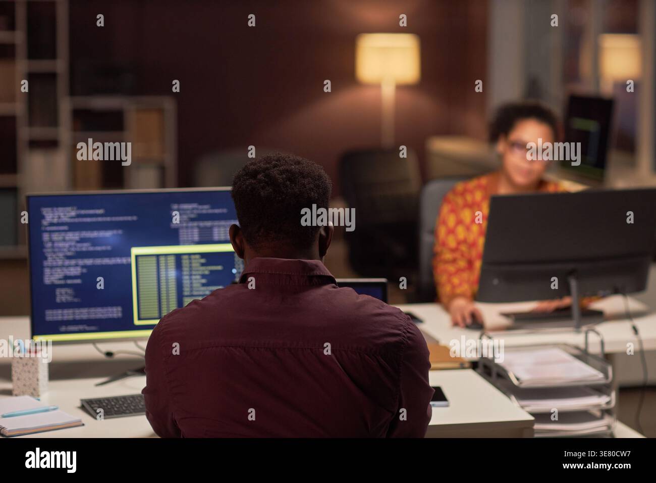 Black male programmer examining computer codes while sitting at table near mixed-race female colleague working late at night in office Stock Photo