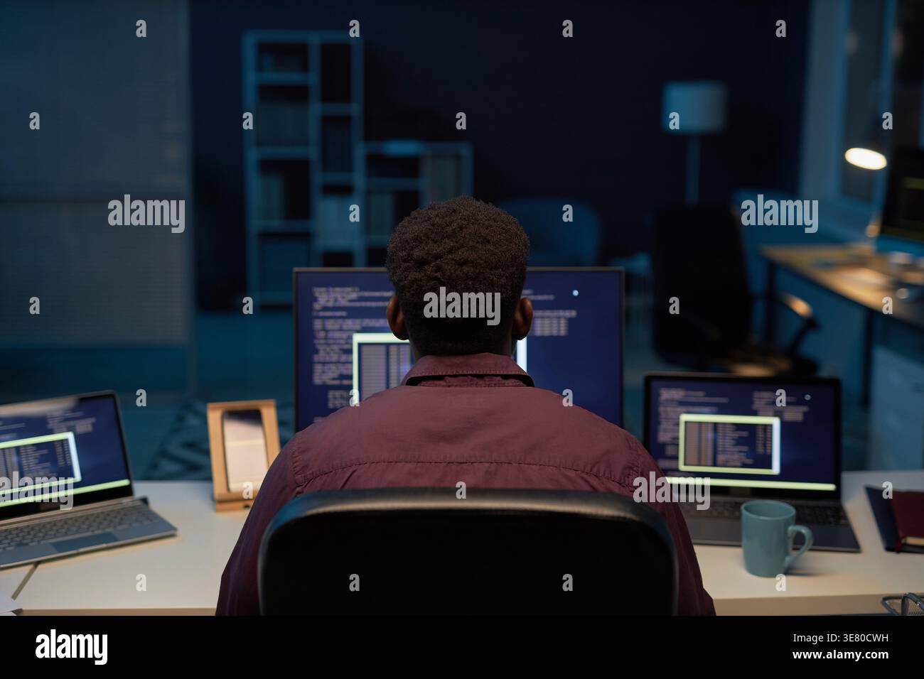 Black male programmer typing computer codes while sitting at table with laptops working late at night in office Stock Photo