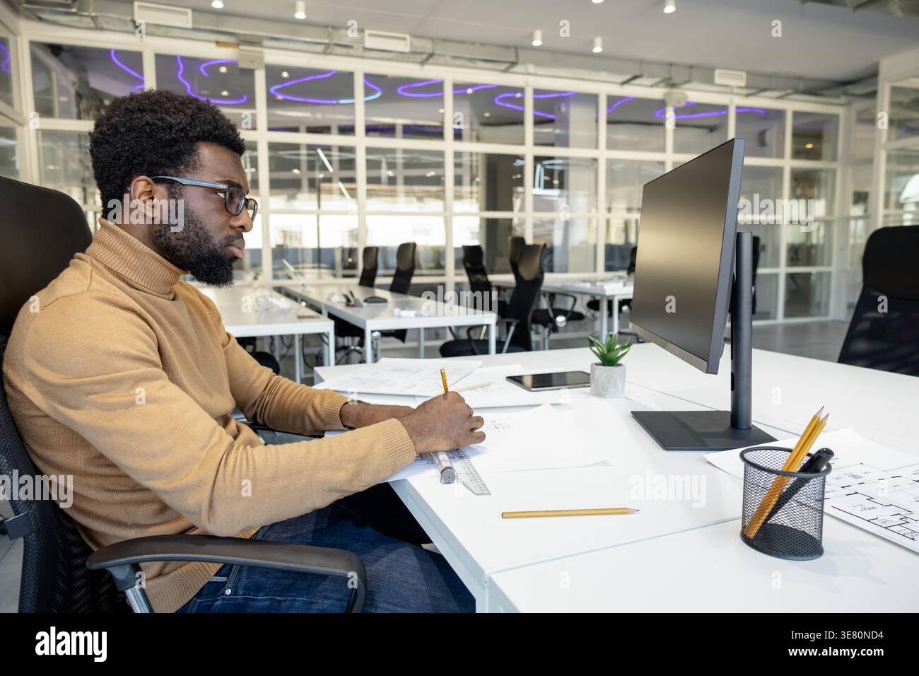 Black male architect with beard wearing brown turtleneck and eyeglasses working on blueprints for new project while sitting at desk with computer i... Stock Photo