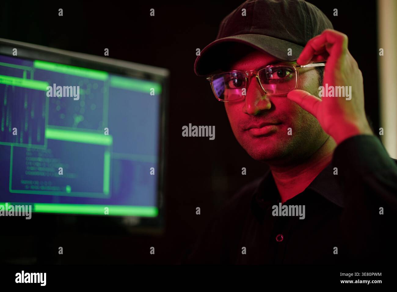 Indian male hacker adjusting glasses sitting at table with computer in security office Stock Photo