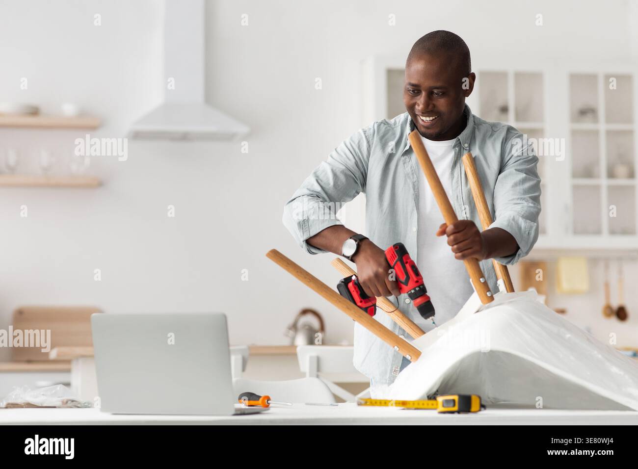 Black man using drill assembling chair on table standing in kitchen at home Stock Photo