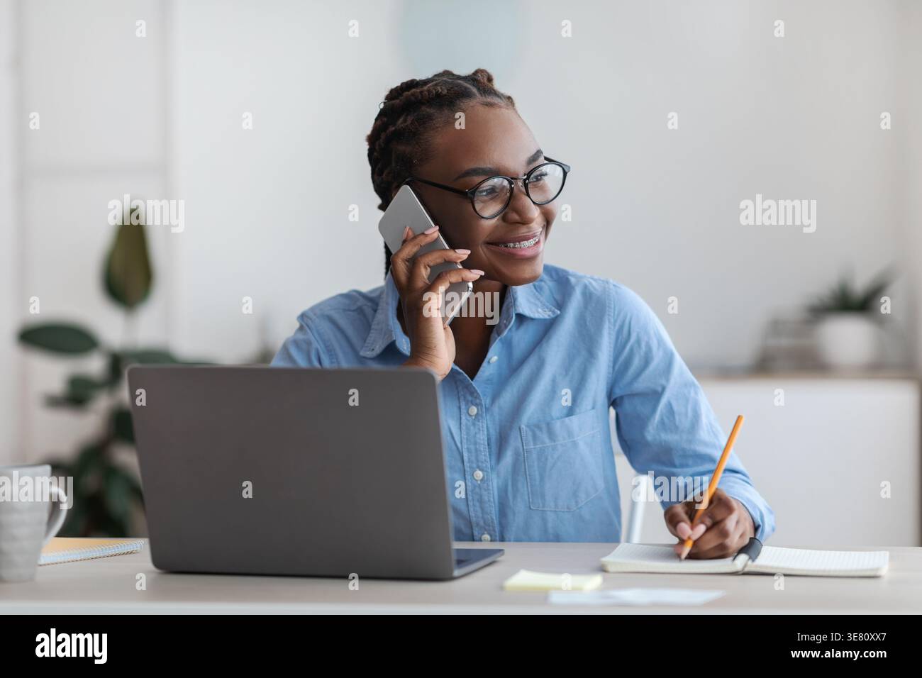 Black business woman with braids wearing eyeglasses having phone call taking notes while sitting at table with laptop in home office Stock Photo