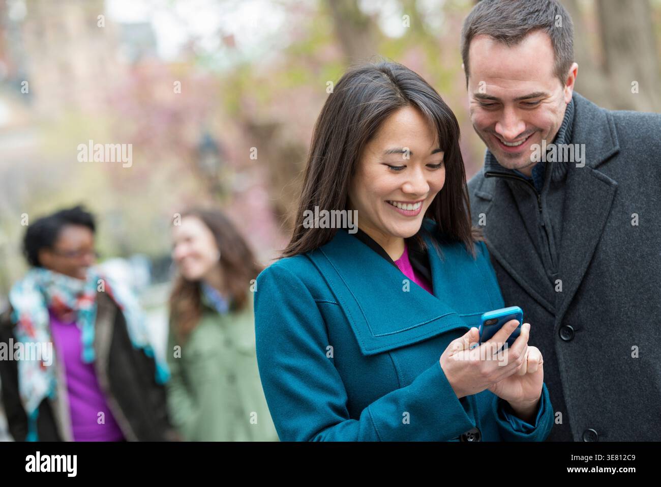 Korean woman showing smartphone to caucasian male friend standing in park during daytime Stock Photo
