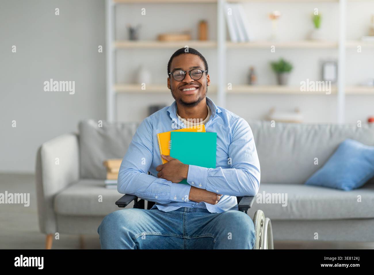 Smiling Black disabled man wearing eyeglasses embracing notebook while sitting on wheelchair in living room at home Stock Photo