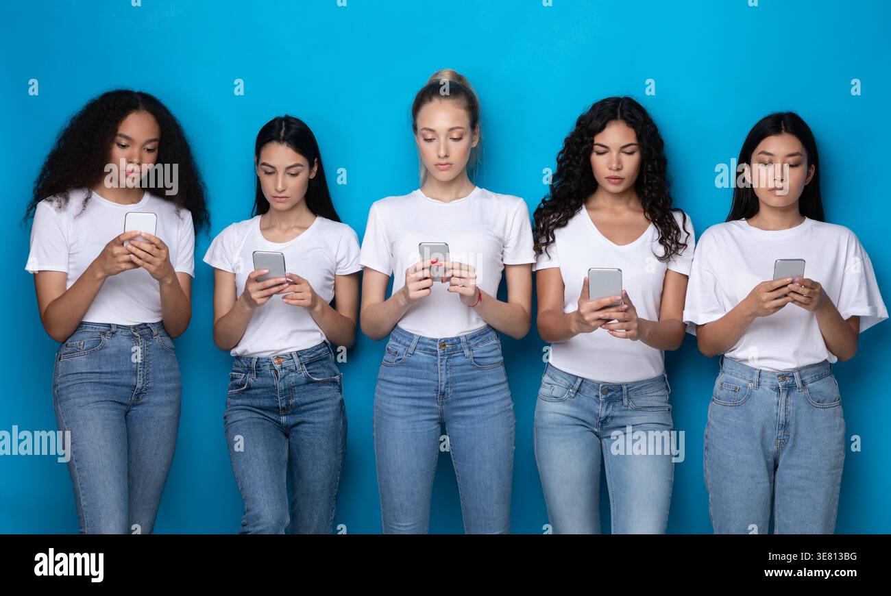 Diverse group of women wearing white shirts and jeans using smartphones standing against blue background in studio Stock Photo