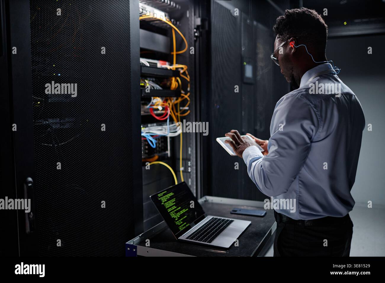 Black male network engineer using tablet and laptop while standing and setting up servers in data center Stock Photo