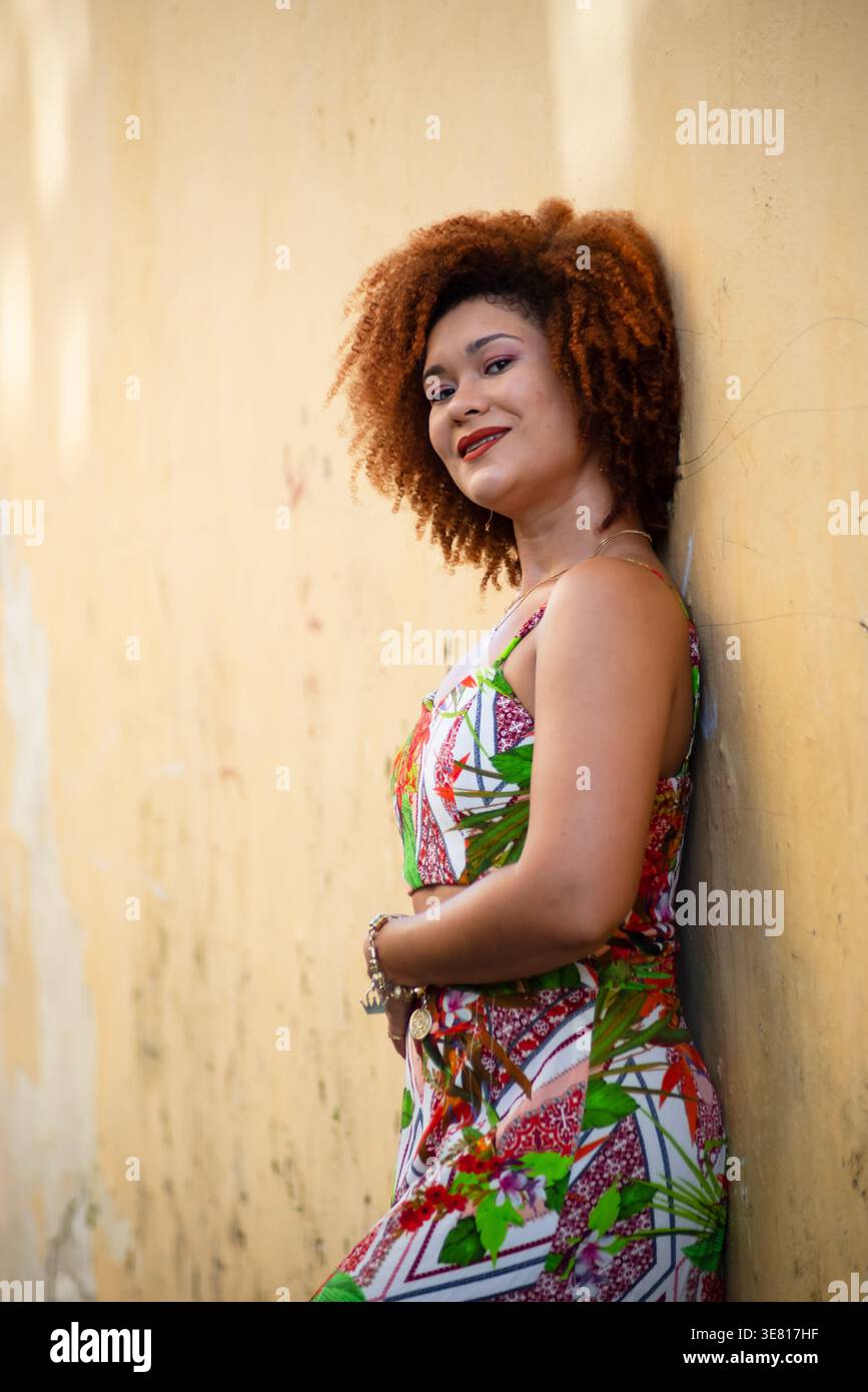 Smiling Black woman with curly afro wearing dress leaning against wall outdoors Stock Photo