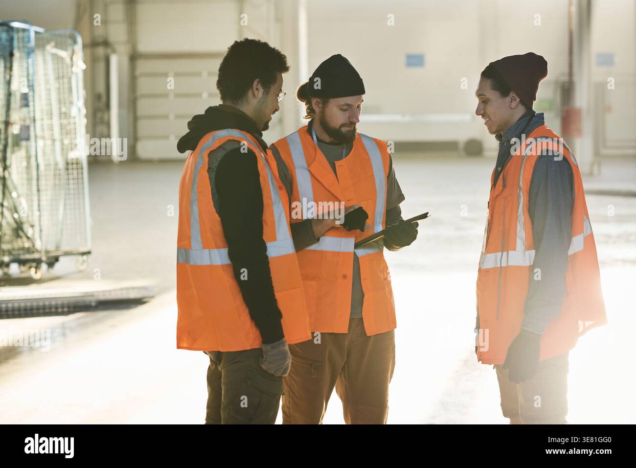 Mixed-race male workers wearing safety vests looking at caucasian male colleague holding tablet standing in warehouse Stock Photo