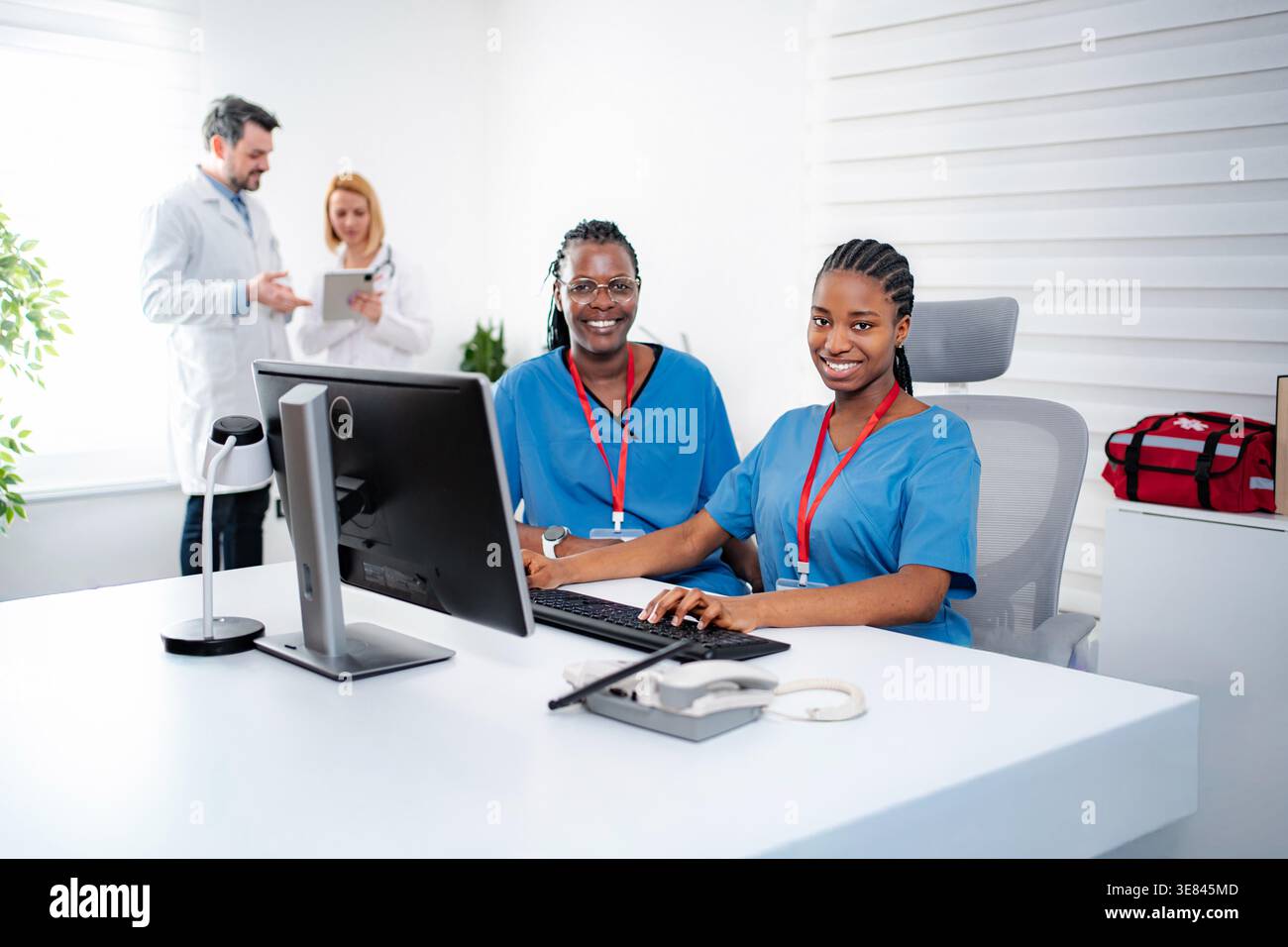 Two smiling nurses using a computer with doctors discussing in the background Stock Photo