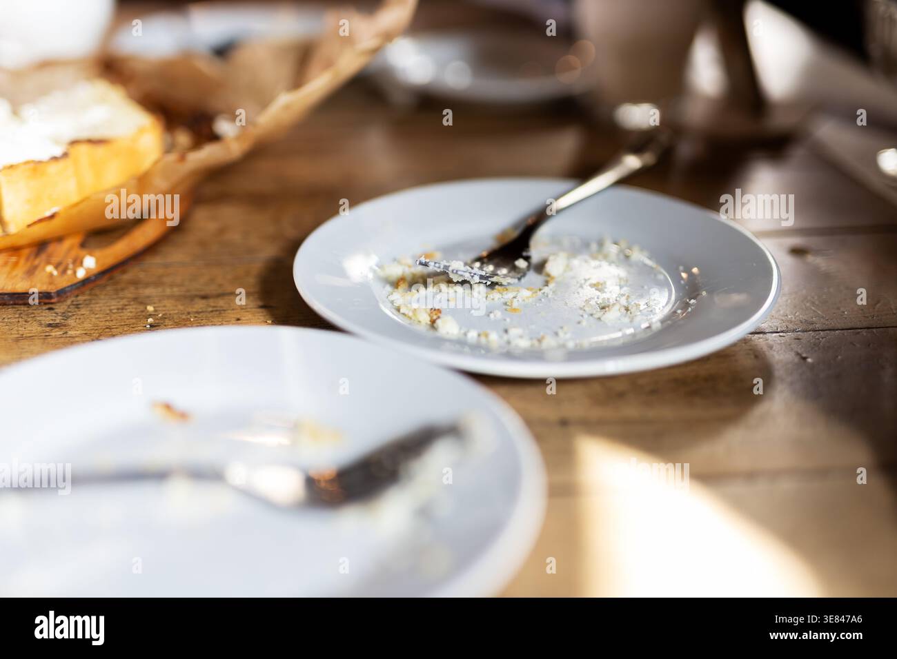 Sunlit breakfast aftermath on wooden table showing empty plates with crumbs, spoon and fork, basket with bread in soft Stock Photo