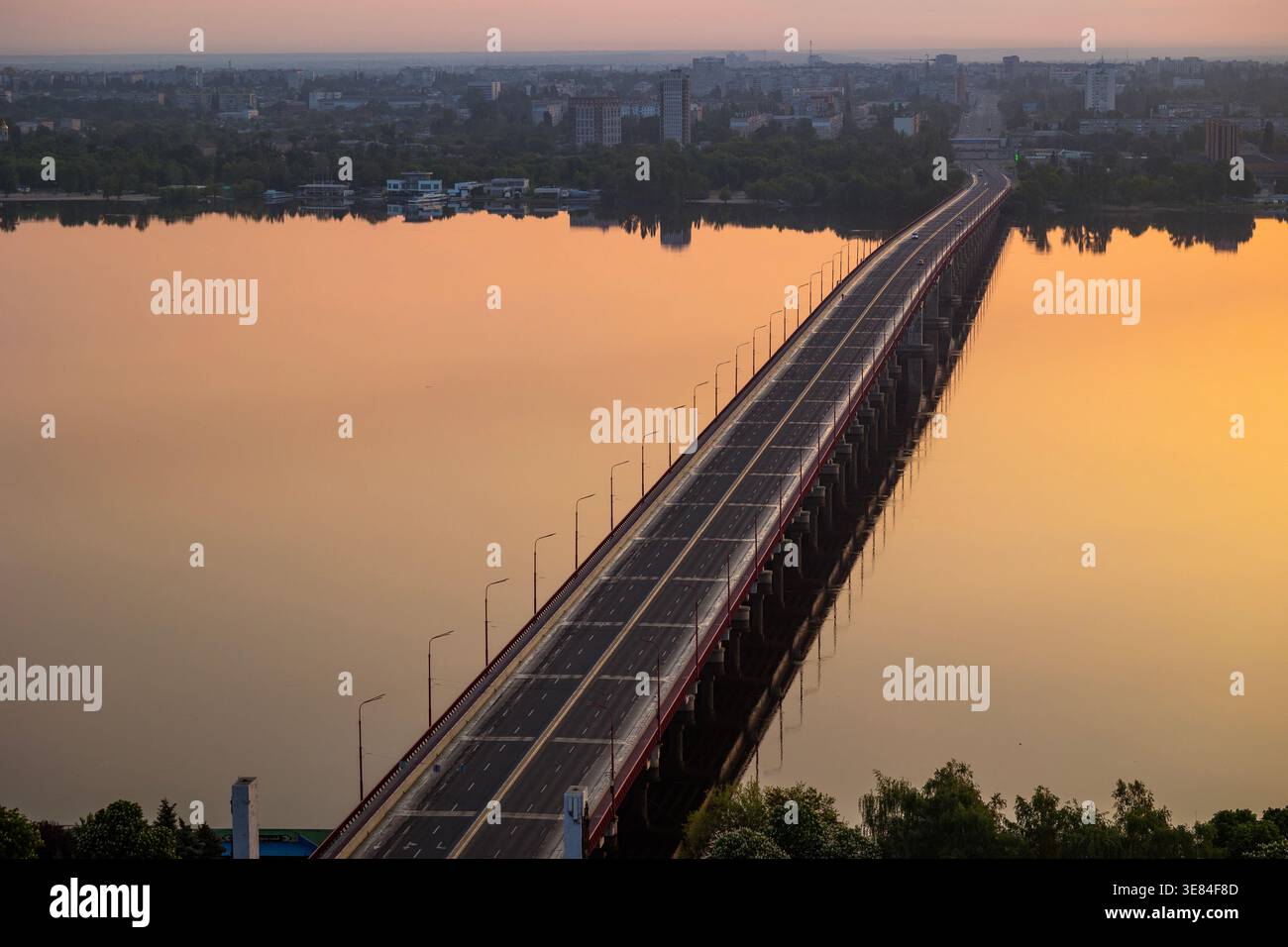 Dnipro, Ukraine - A peaceful sunrise or sunset bathes a long, wide multilane bridge and the surrounding urban landscape in warm, golden hues, reflecti Stock Photo