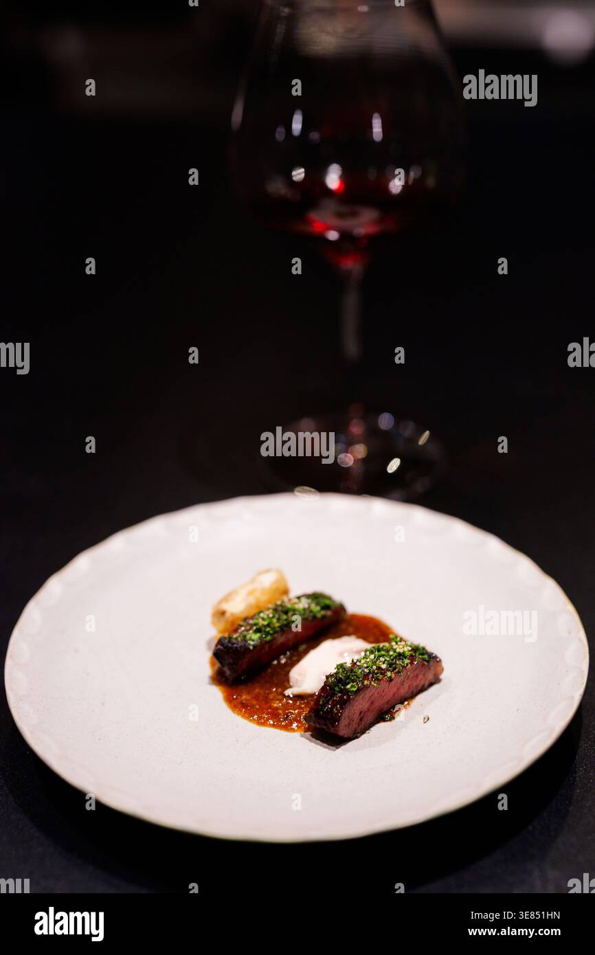 Gourmet plated beef medallions with herb crust, sauce and puree on white plate, moody fine dining setting with red wine glass bokeh. Stock Photo
