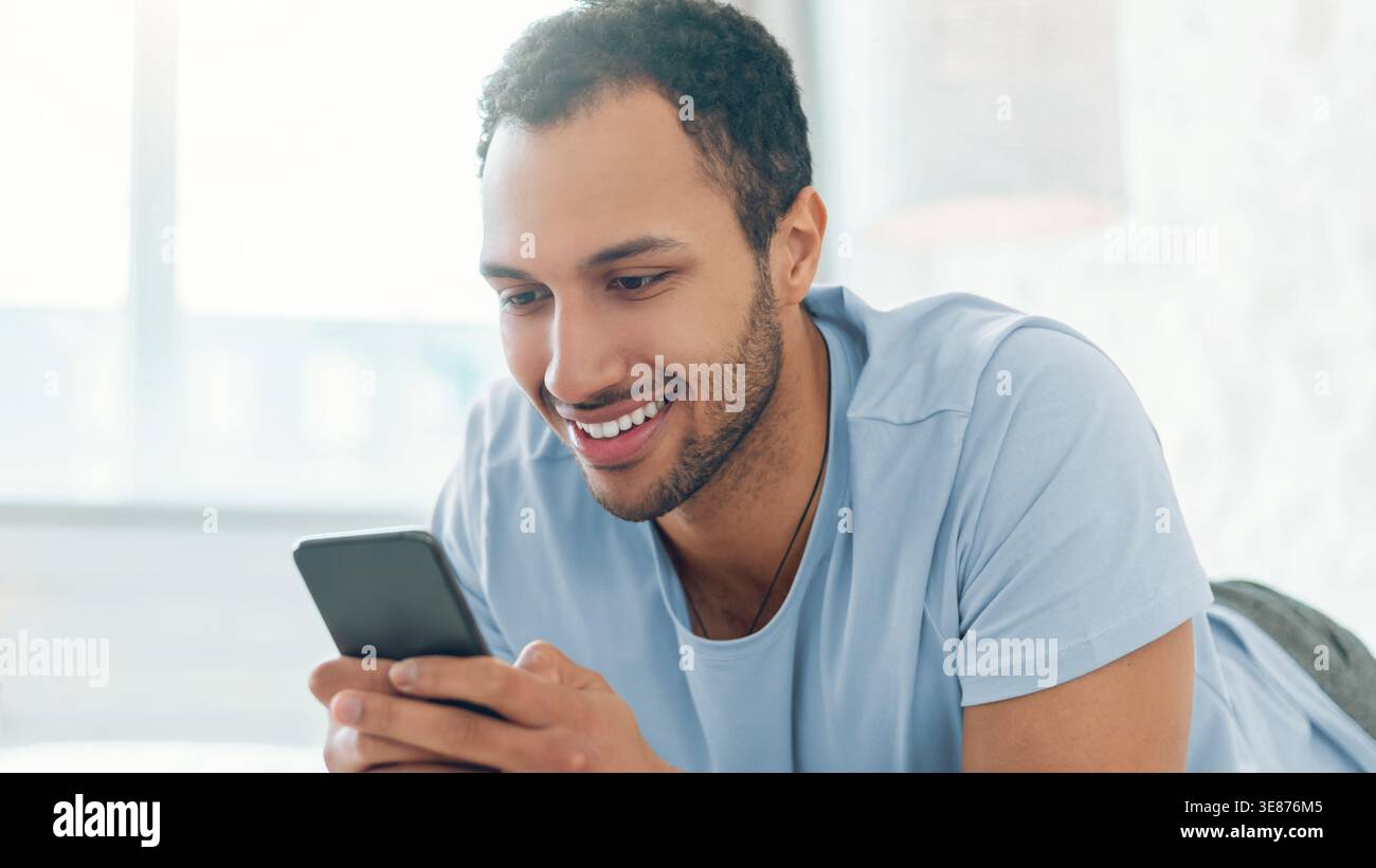Young Man Smiles While Using Smartphone at Home During Daytime Activity Stock Photo
