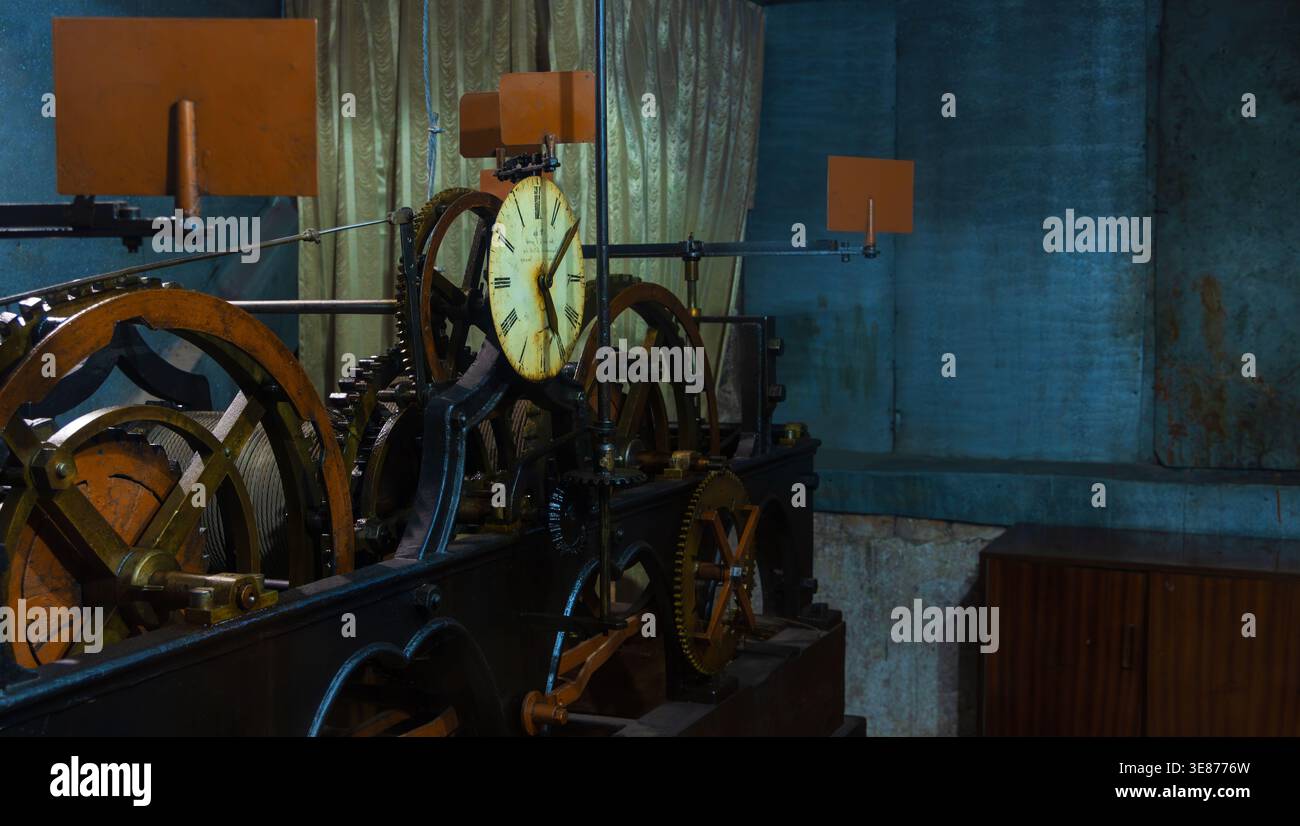 A vintage mechanical clock with a large yellow face and black hands is surrounded by rusty metal gears and cogs in a dimly lit room with blue walls, a Stock Photo