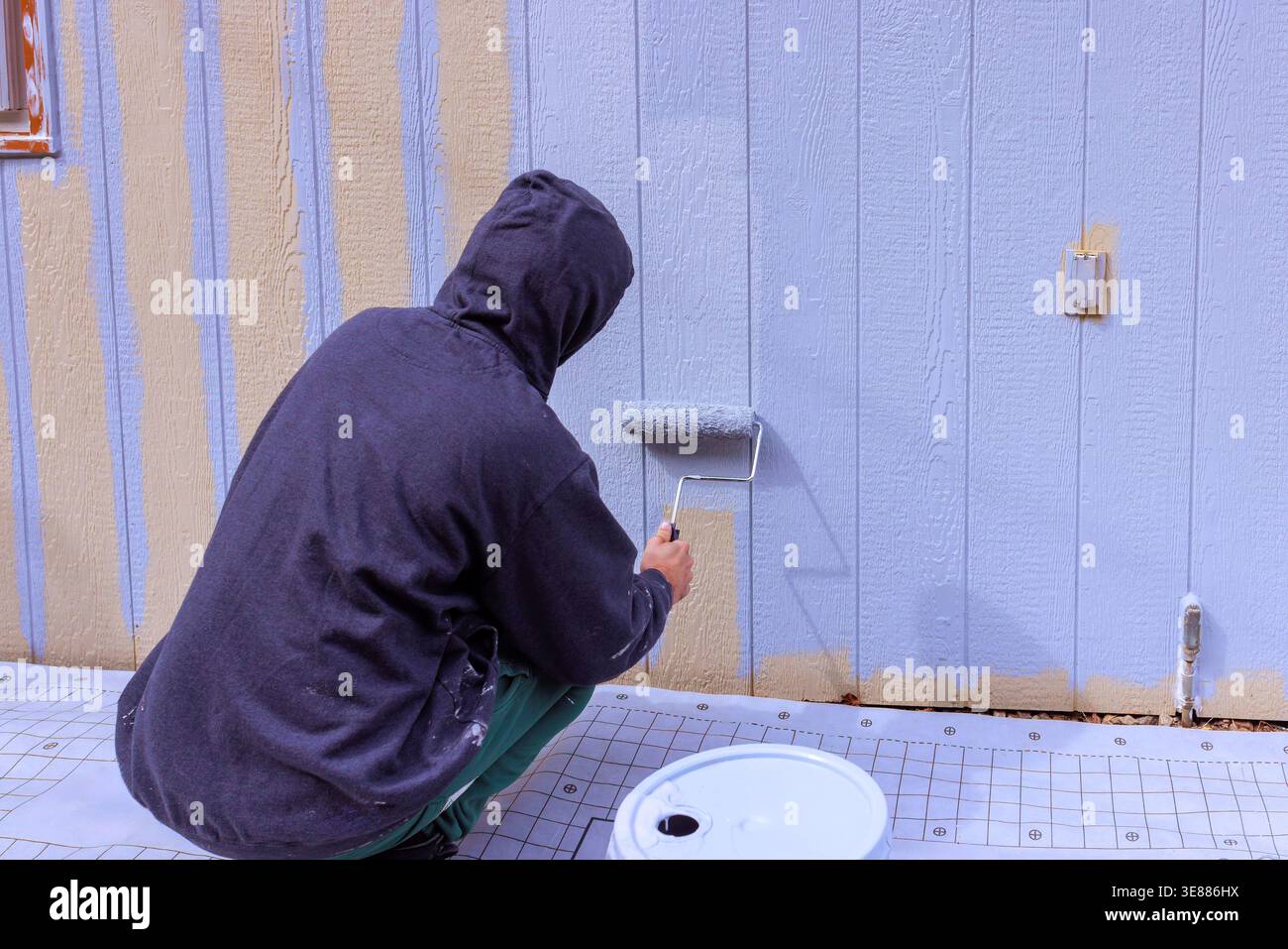 Someone is applying paint to siding panel wall using roller while sitting on ground in construction setting. Stock Photo