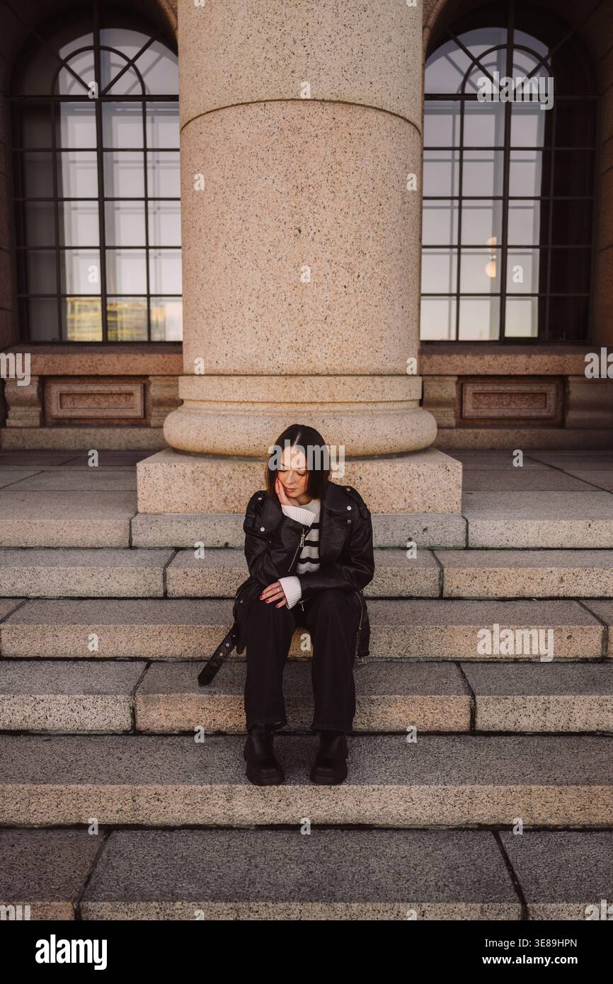 Stylish woman in leather jacket sitting on stone steps in city Stock Photo