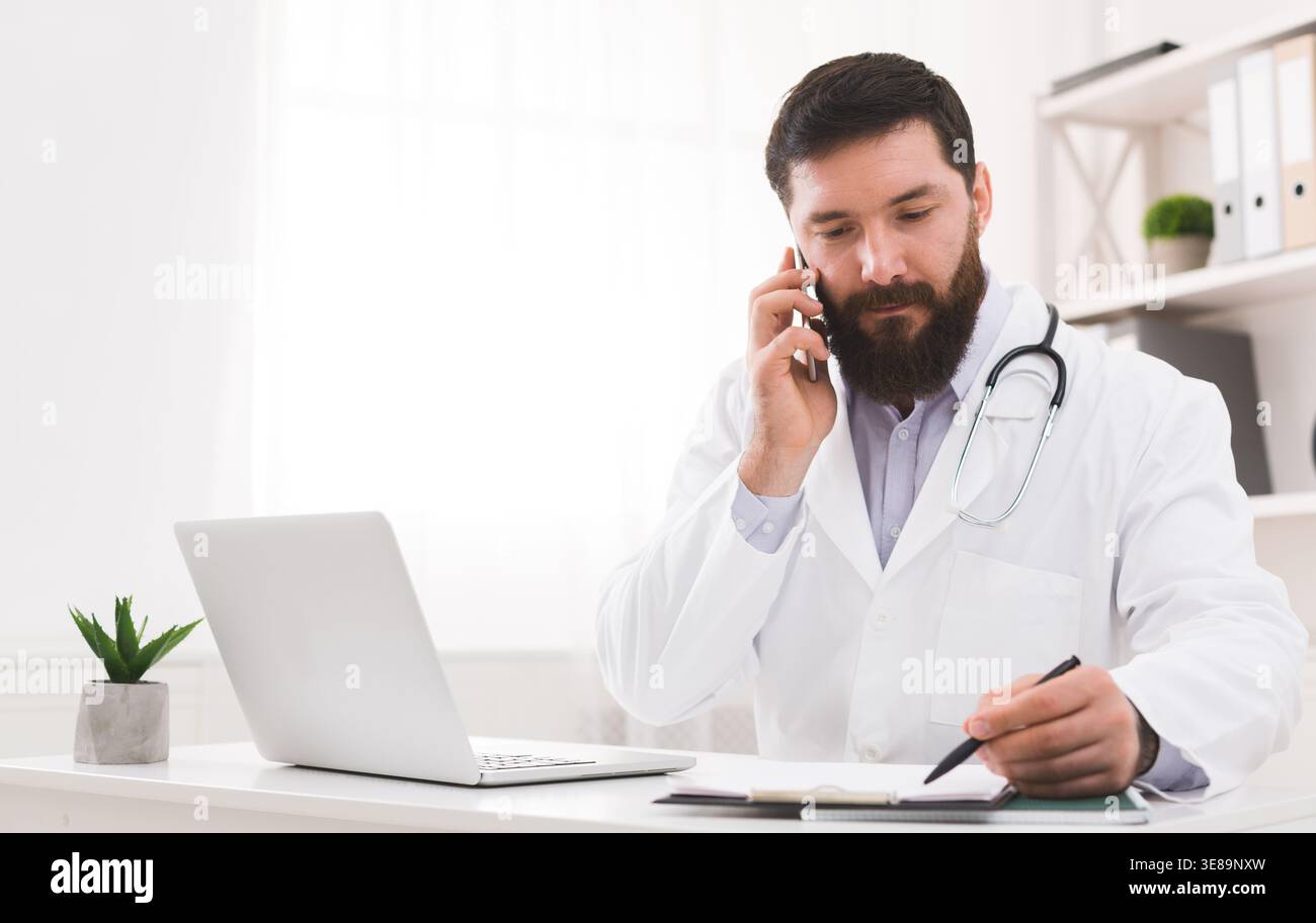 Doctor on the Phone While Taking Notes at the Desk in His Office During Daytime Stock Photo