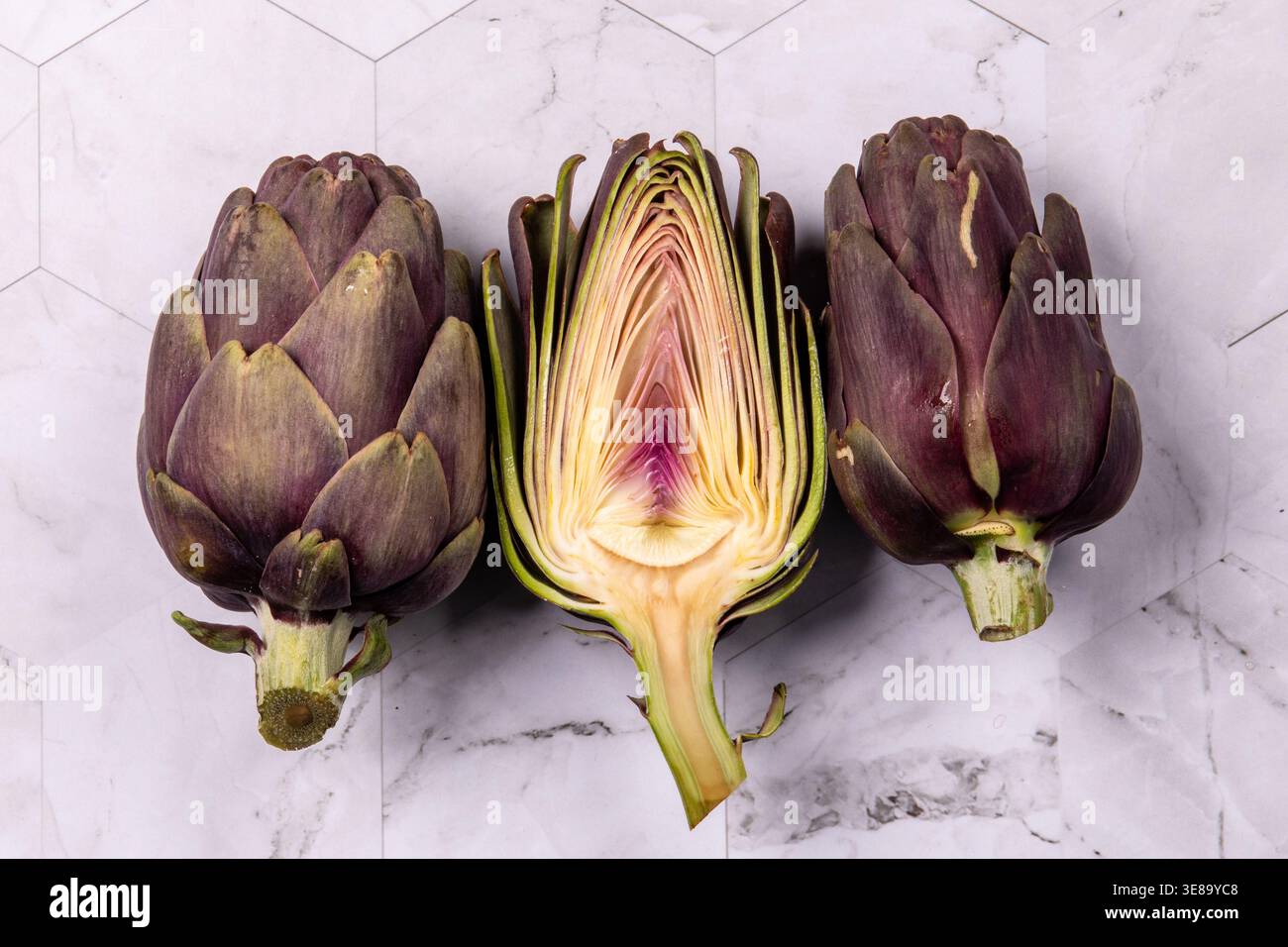 Three artichokes placed on a marble surface, one sliced. Stock Photo