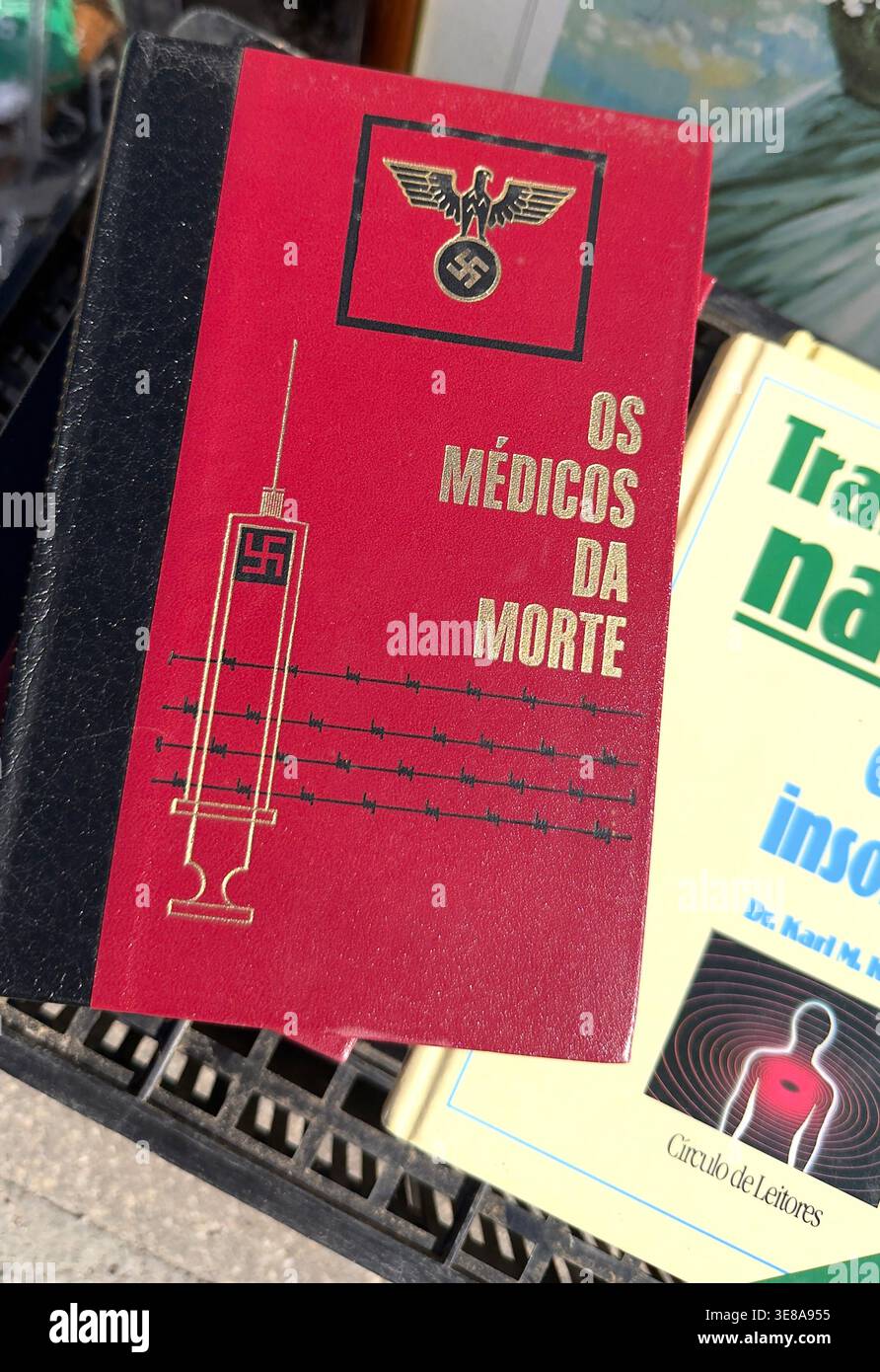 Portuguese book titled Os Medicos da Morte featuring a red cover with Nazi eagle swastika and syringe illustration on a plastic crate - Smartphone Captured Stock Image