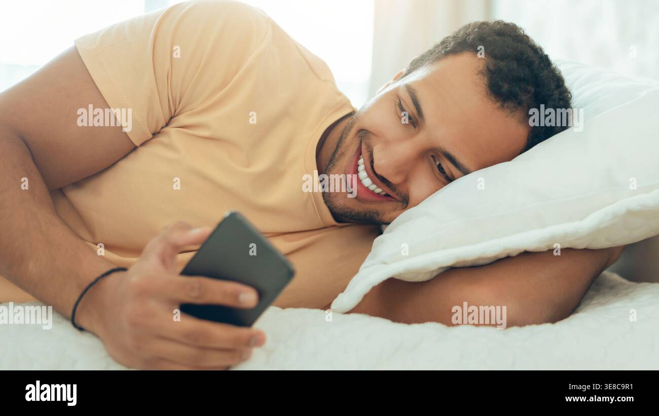 Man Lying on Bed Using Smartphone and Smiling While Looking at Screen During Daytime Stock Photo
