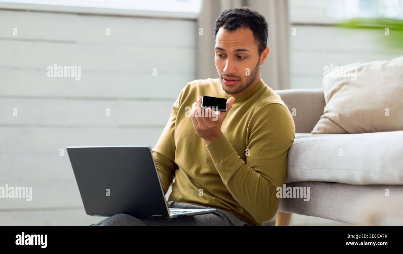 Man Using Voice Command on Smartphone While Working on Laptop in Living Room Space Stock Photo