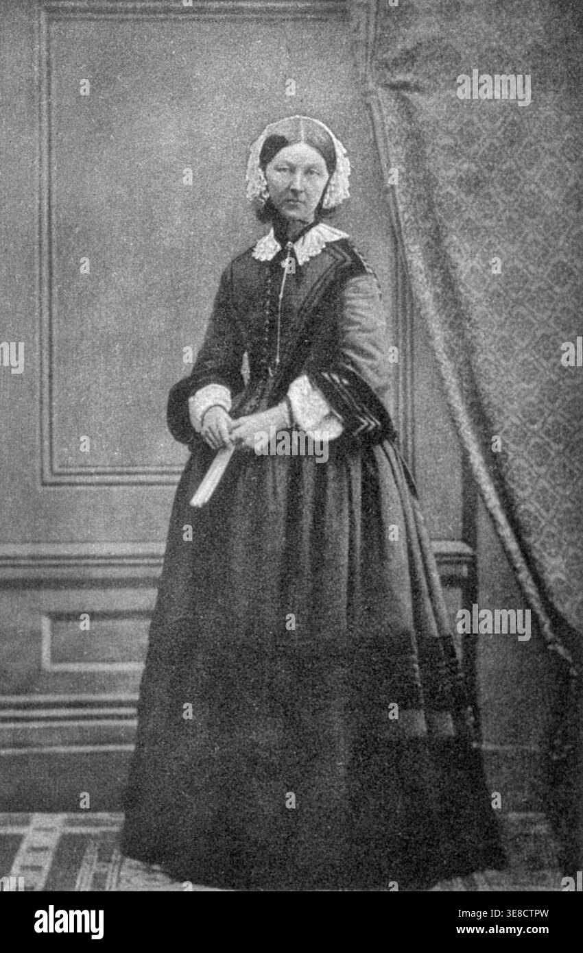 Photogravure of Florence Nightingale by E. Walker after Goodman, 1858. She  stands full-length wearing a dark dress with a lace collar and white cap,  positioned in a Victorian interior beside a draped