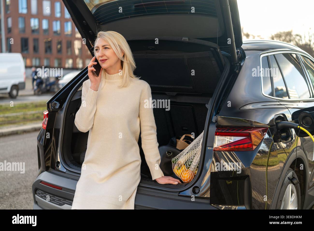 A professional woman speaking on her mobile phone while standing next to the open trunk of an electric vehicle in an urban environment. Stock Photo