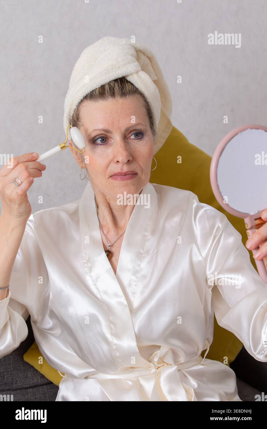 A woman dressed in a silk robe is attentively applying skincare using a device while seated in a comfortable chair. She looks into a handheld mirror, Stock Photo