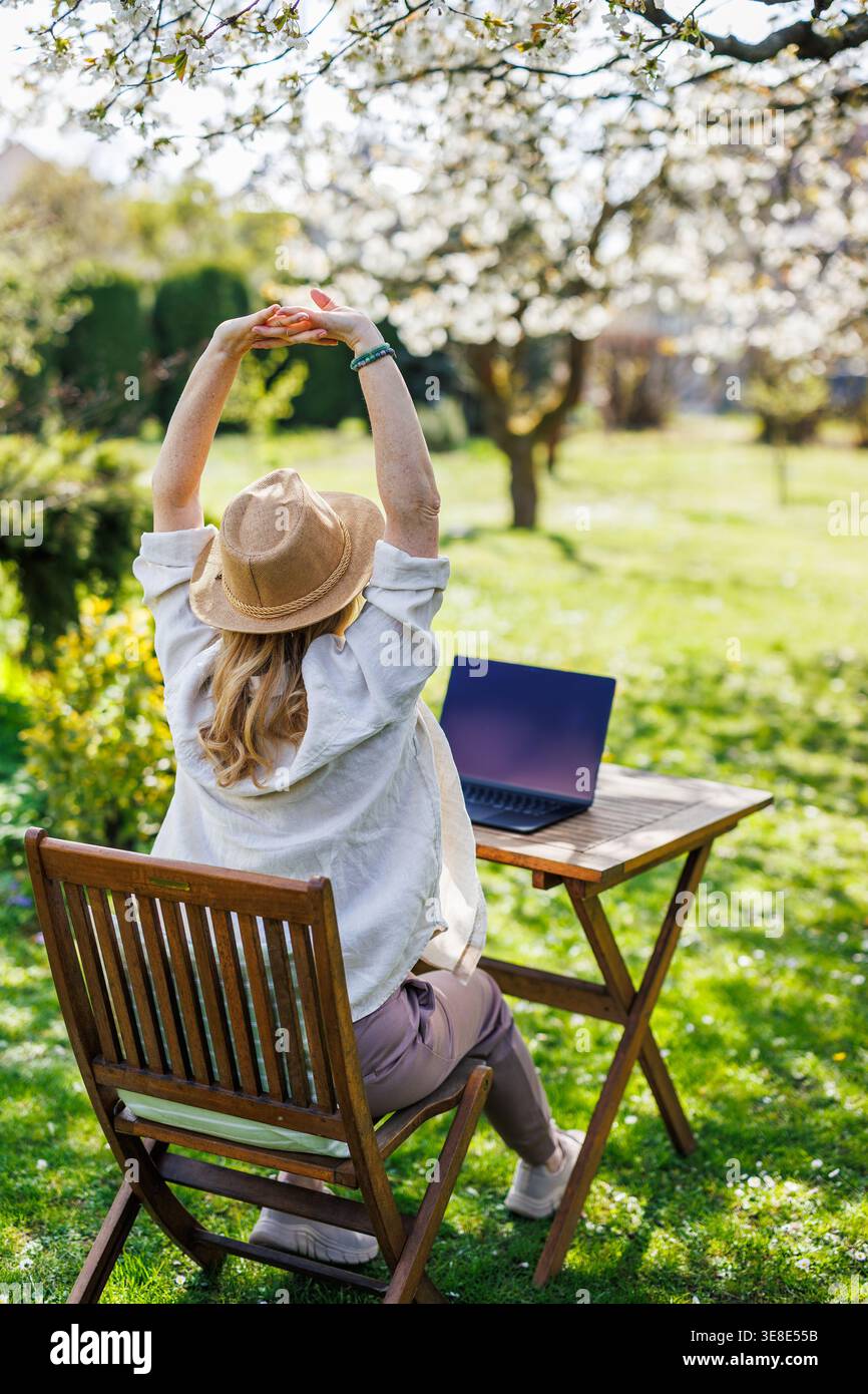 Woman stretching hands while working with laptop outdoors. Stress relief. Exercising body during remote work break in spring garden Stock Photo