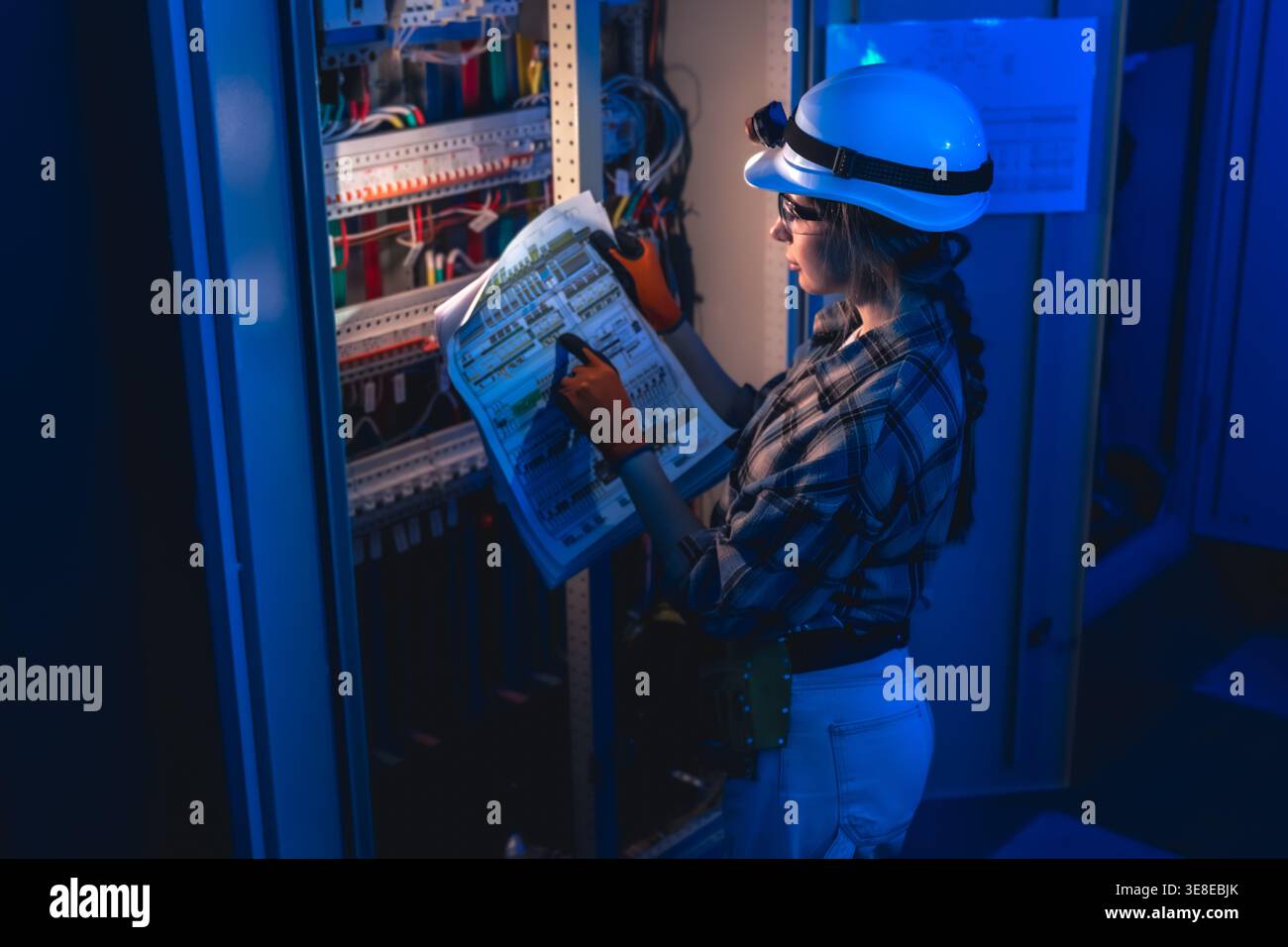 Electrician inspecting electrical switchboard using technical blueprint. Industrial energy system. Stock Photo