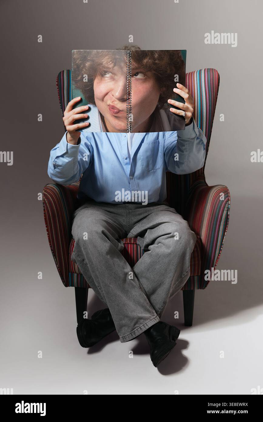 Young man sitting in armchair holding notebook covering face minimal studio portrait. Stock Photo