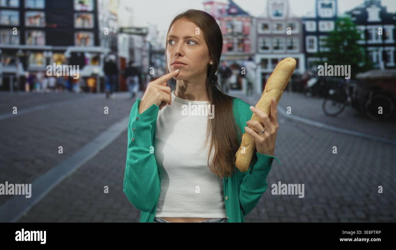 Woman holding a baguette with finger to chin on city street lined with buildings and bicycles; thoughtful reflection. Stock Photo