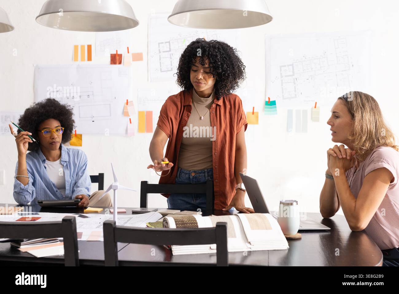 Diverse female coworkers studying blueprints at design studio, standing woman holding yellow object Stock Photo