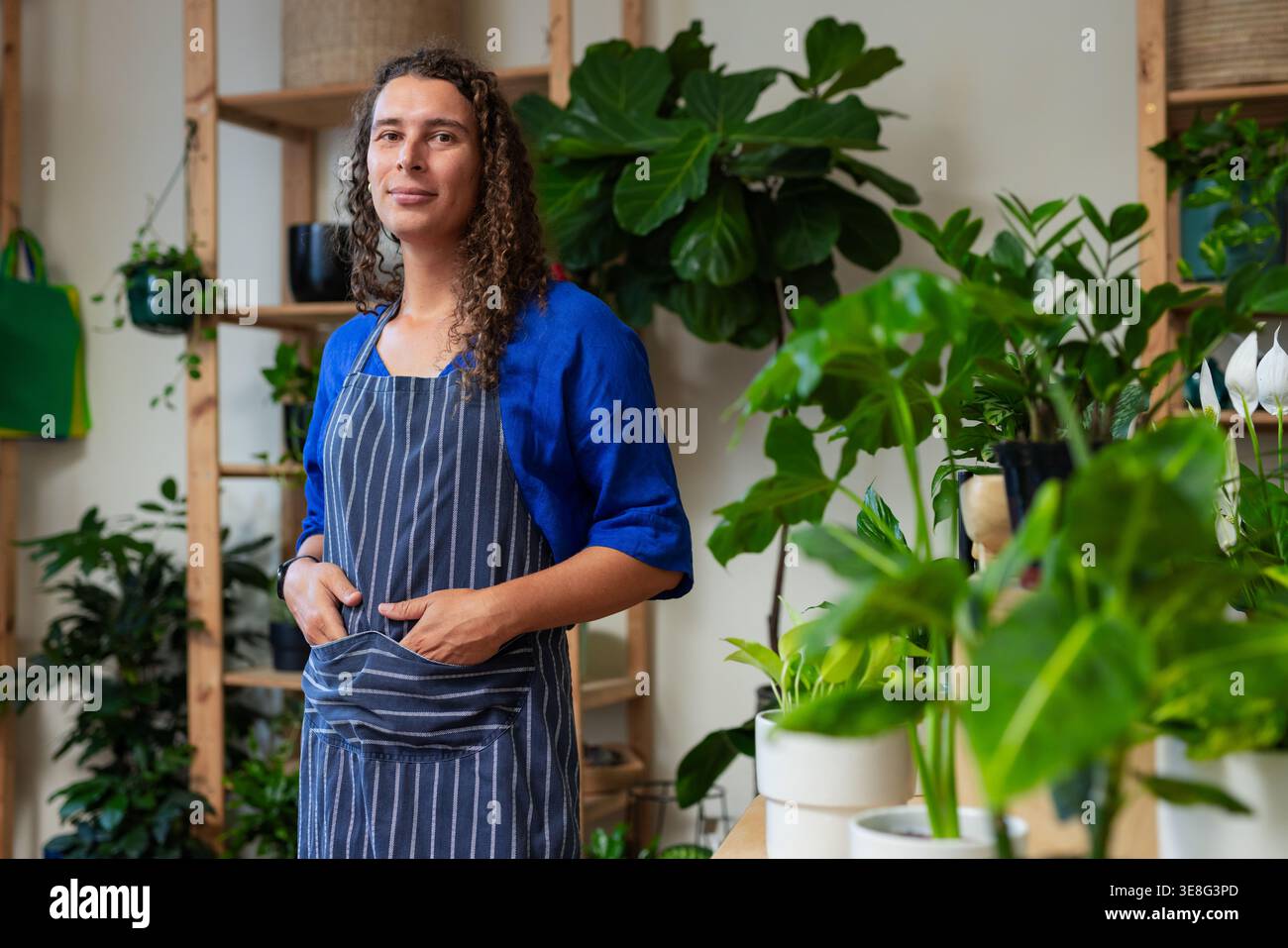 Non-binary shop worker arranging pots and watering houseplants on work counter in striped apron Stock Photo