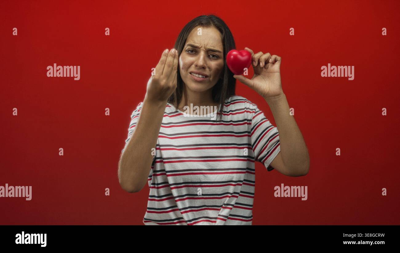 Young hispanic woman holds red heart near face and beckons with hand in studio; pleading love appeal. Stock Photo