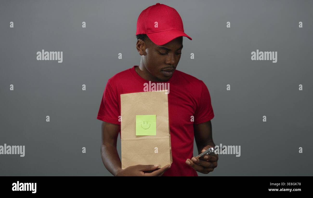 Young man holding smartphone and paper bag with smiley note while looking down in gray studio; curiosity. Stock Photo