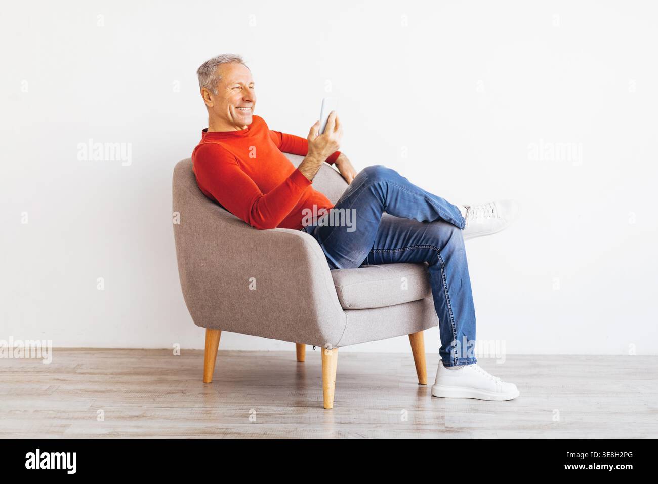Older Man Sits on Chair and Smiles While Using Smartphone in Indoor Setting Stock Photo