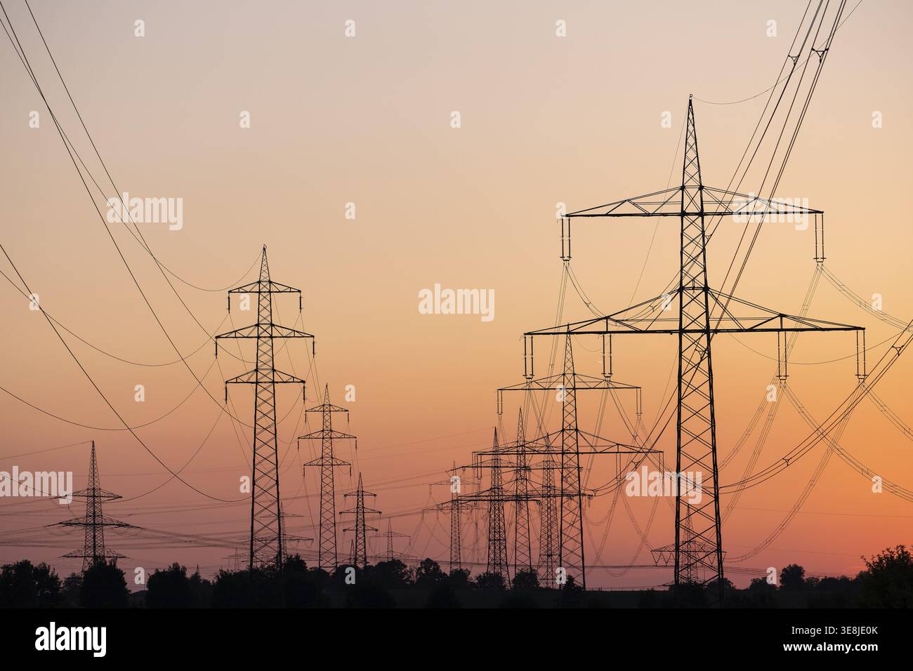 Silhouette of several high-voltage power lines and electricity pylons against a glowing orange and red sunset sky in Germany Stock Photo