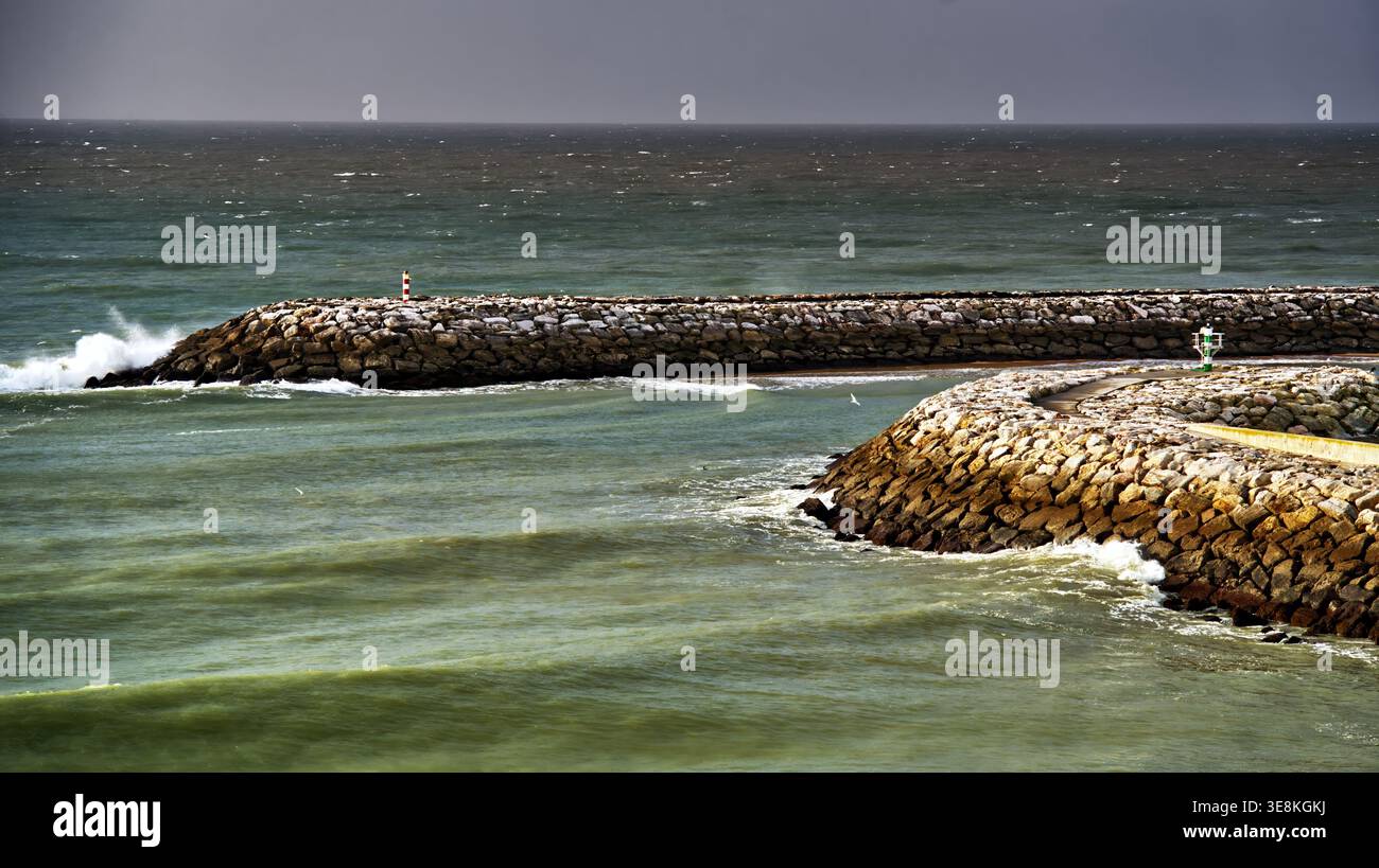 Ocean waves crashing against the rocky breakwaters of albufeira marina, with a small lighthouse marking the entrance under a brooding sky in portugal' Stock Photo