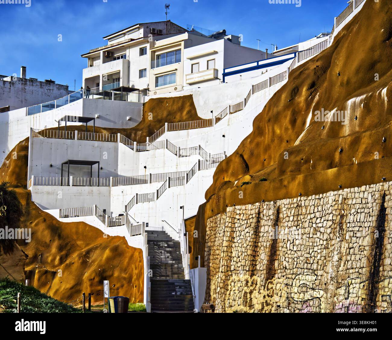 White modern buildings and long concrete staircases climb a steep terracotta cliff in albufeira, portugal, under a bright blue mediterranean sky for a Stock Photo