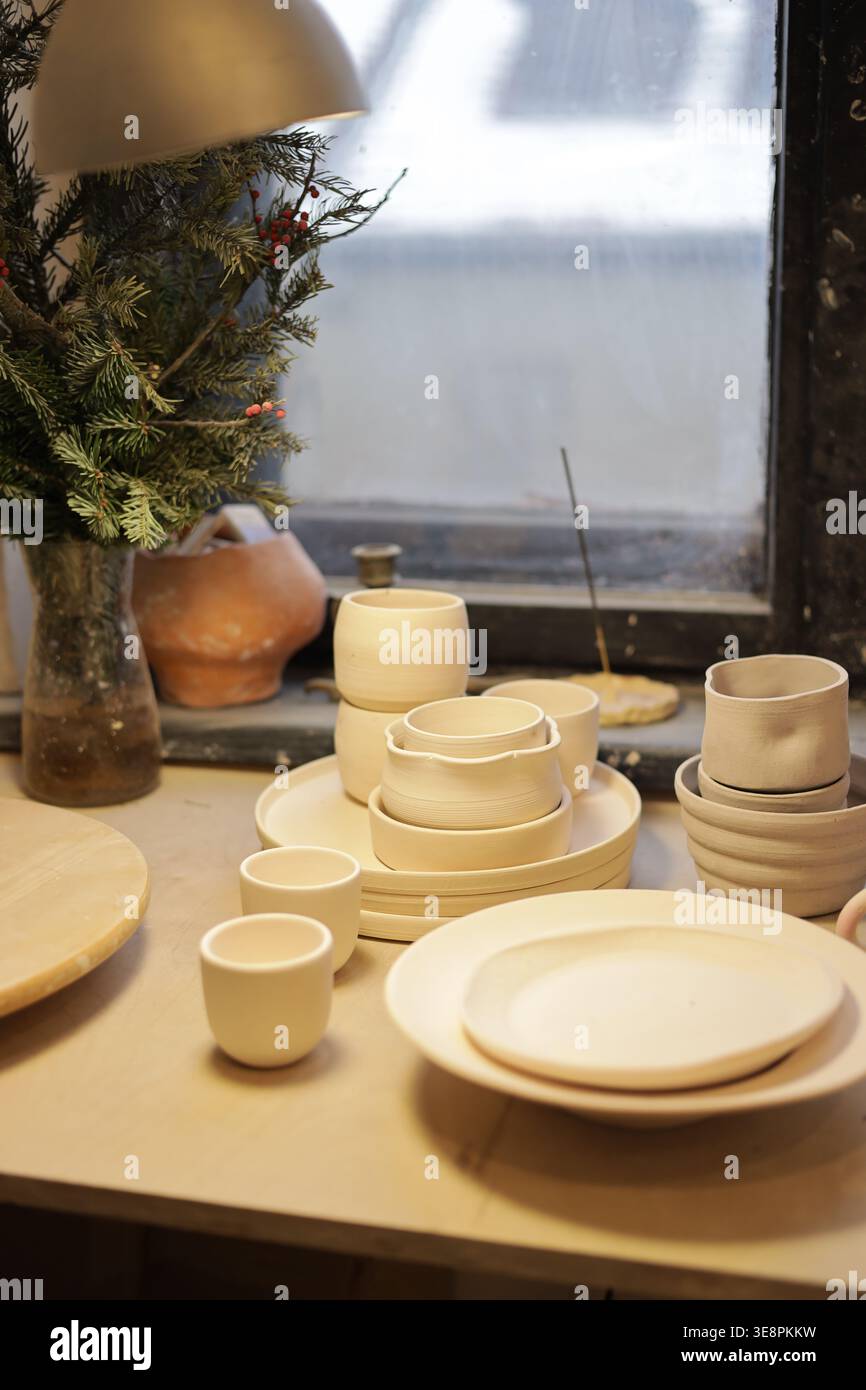 Beautiful still life of a complete handmade ceramic dishware set — plates, cups and bowls — on a bright studio windowsill with a plant. Stock Photo