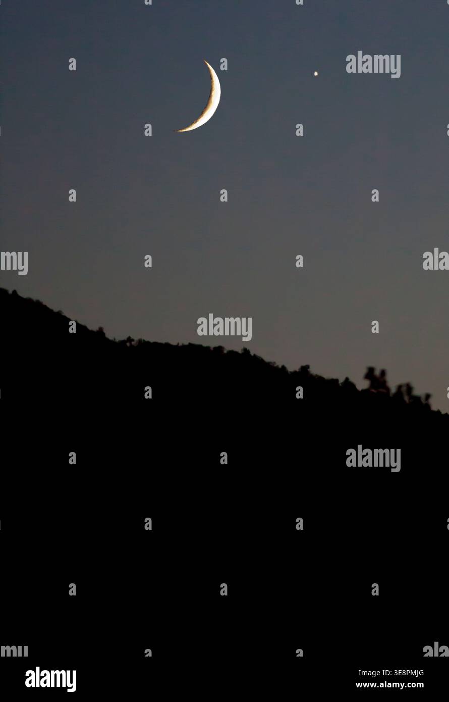 The moon hangs in the sky with a crescent shape while a bright star can be seen close by. The scene captures a dark hill silhouette beneath the night Stock Photo
