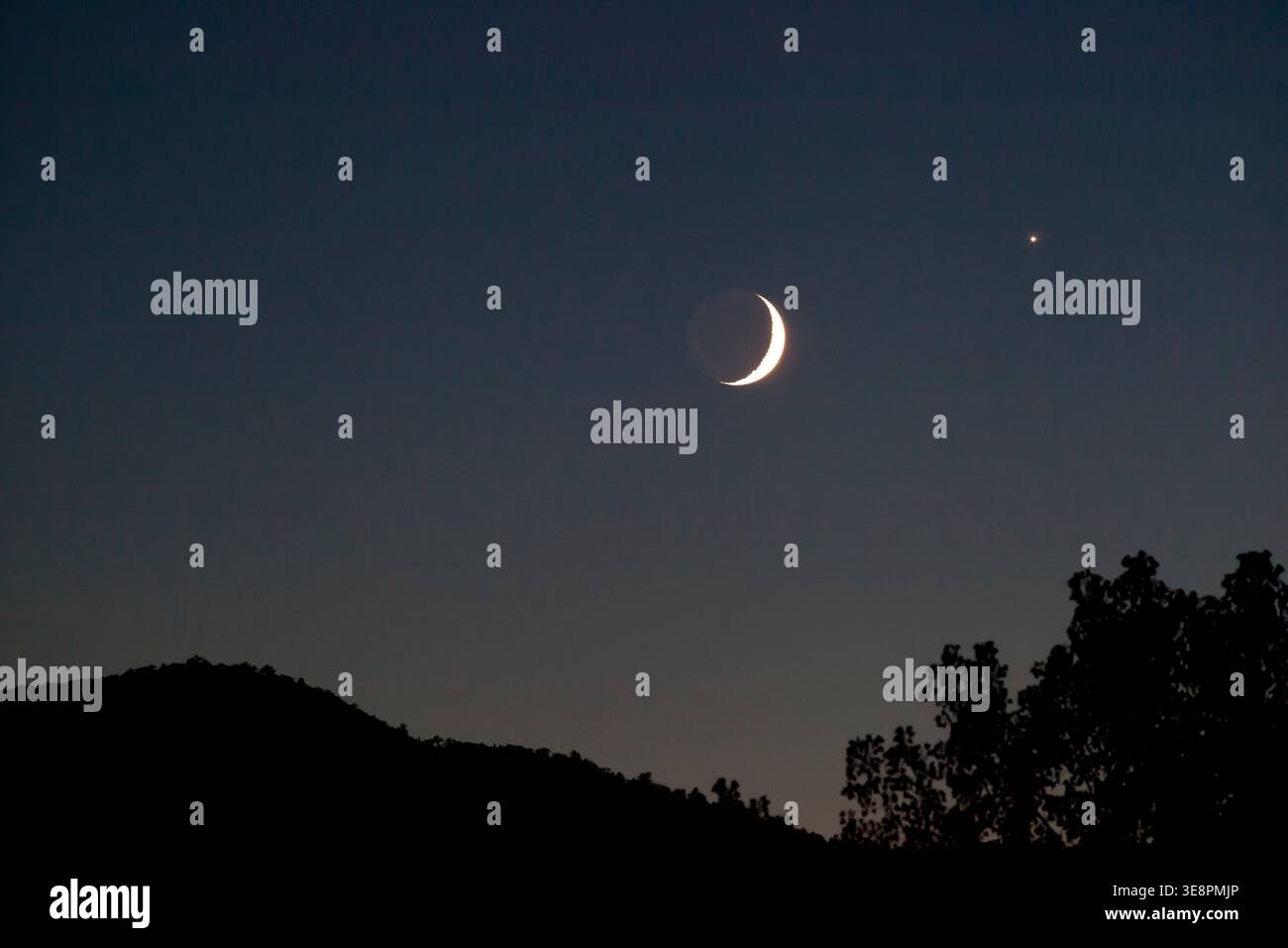 The moon hangs in the sky with a crescent shape while a bright star can be seen close by. The scene captures a dark hill silhouette beneath the night well Stock Photo