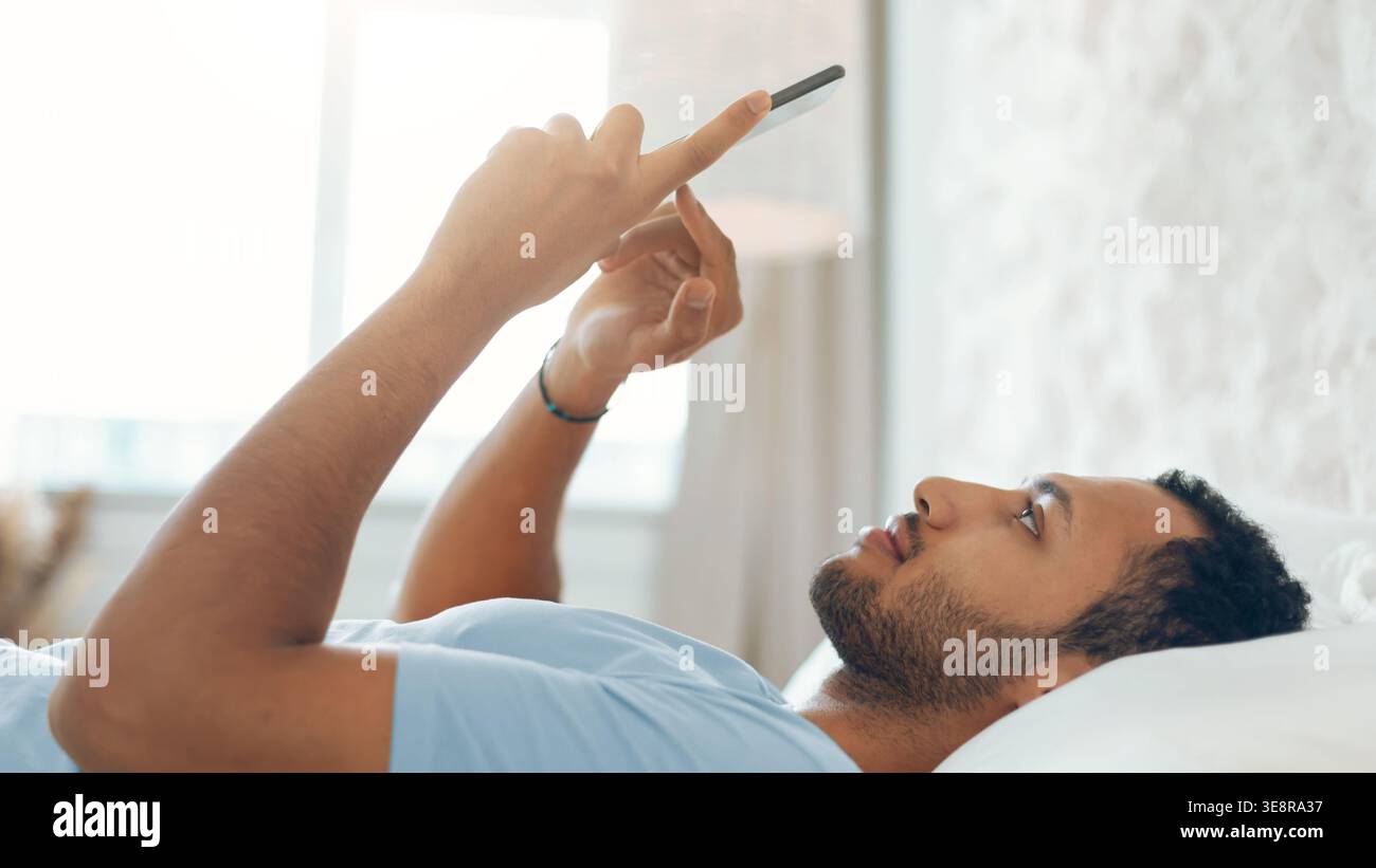 Man Lying on Bed Using Smartphone While Looking at Screen in Bright Room Setting Stock Photo