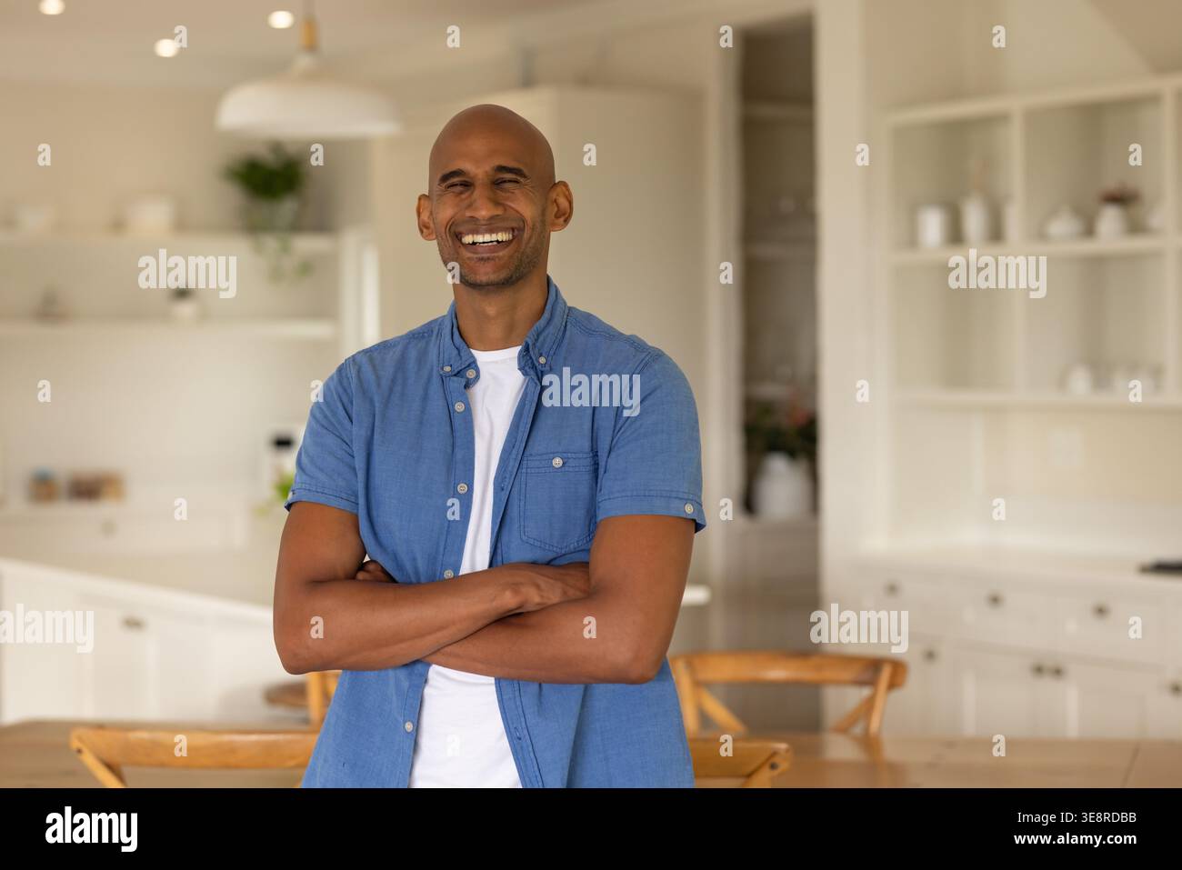 Man standing and smiling wearing blue short-sleeve shirt over white tee in modern kitchen island Stock Photo