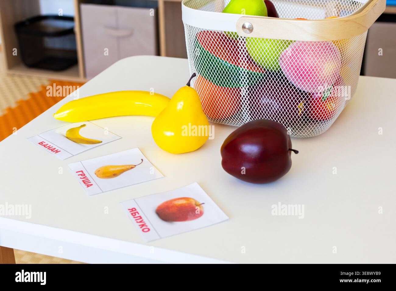 Close view of a basket of toy fruits with banana pear and plum beside word cards on a white table encouraging object to picture matching Stock Photo