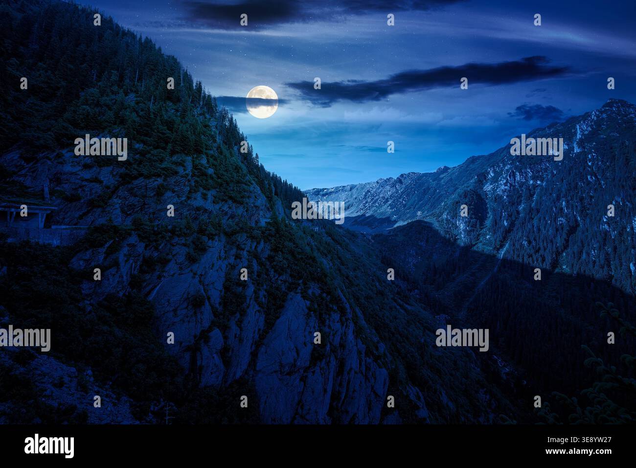 Valley in summer mountains at night. Gorgeous mountainous landscape. full moon over rocky cliffs and forested slopes under dark cloudy sky. Mysterious Stock Photo