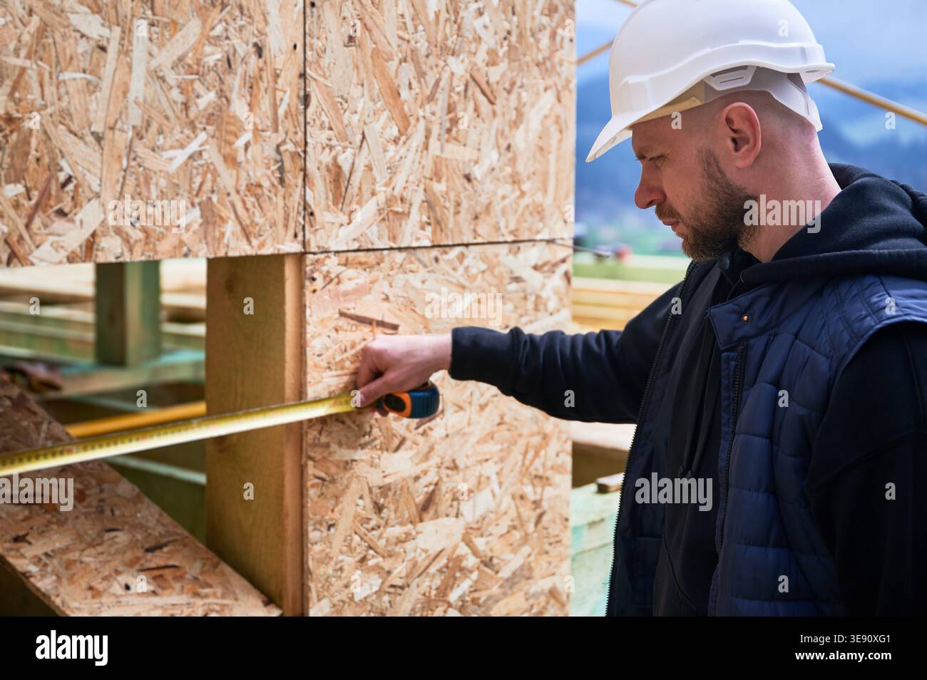Carpenter measuring horizontal distance across wooden facade cutout, verifying alignment and installation accuracy on building site Stock Photo