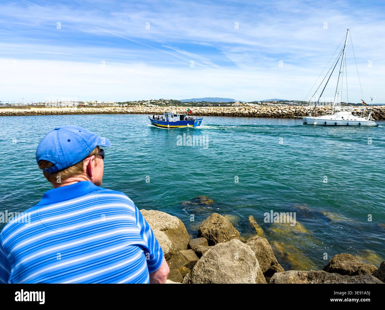 Man in blue striped shirt and baseball cap watching a fishing boat and catamaran enter a harbour in Lagos Algarve Portugal Stock Photo